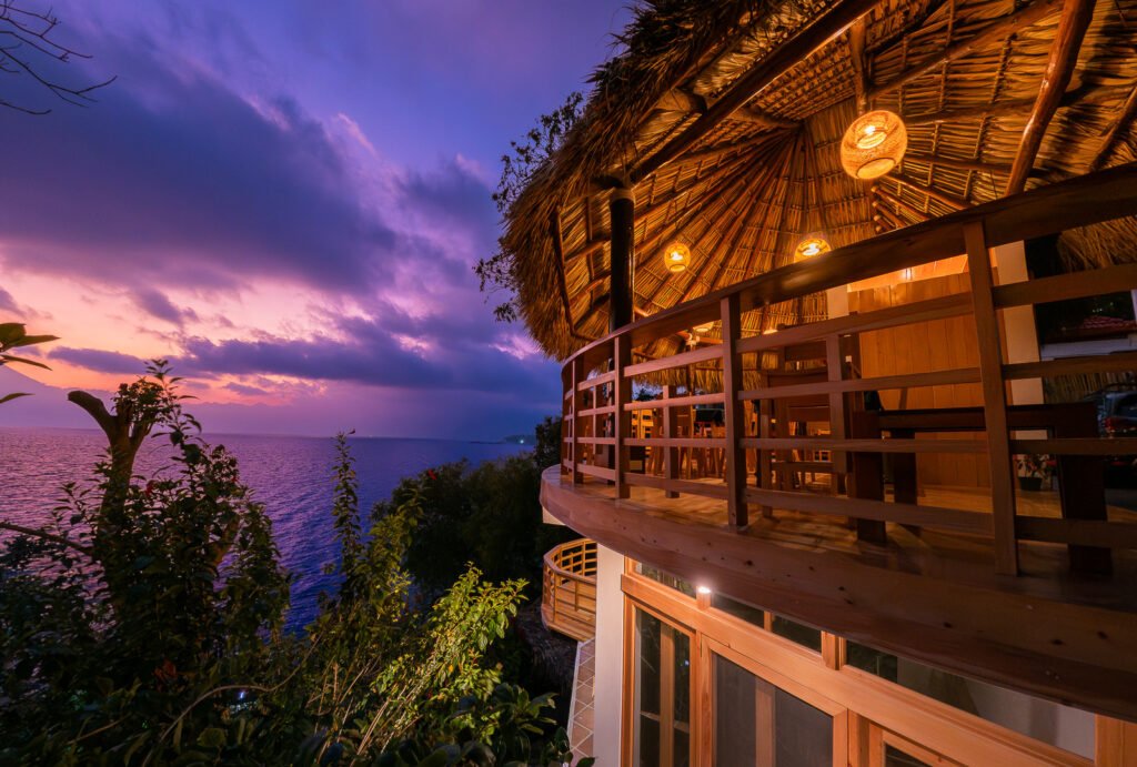 B'alam Ya Cafe balcony view at sunset. Thatched roof, ocean, and purple sky create a serene scene.