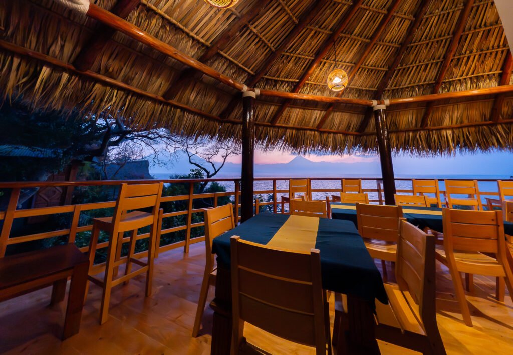 B'alam Ya Cafe view: Tables under a thatched roof overlook a lake and volcano at dusk.