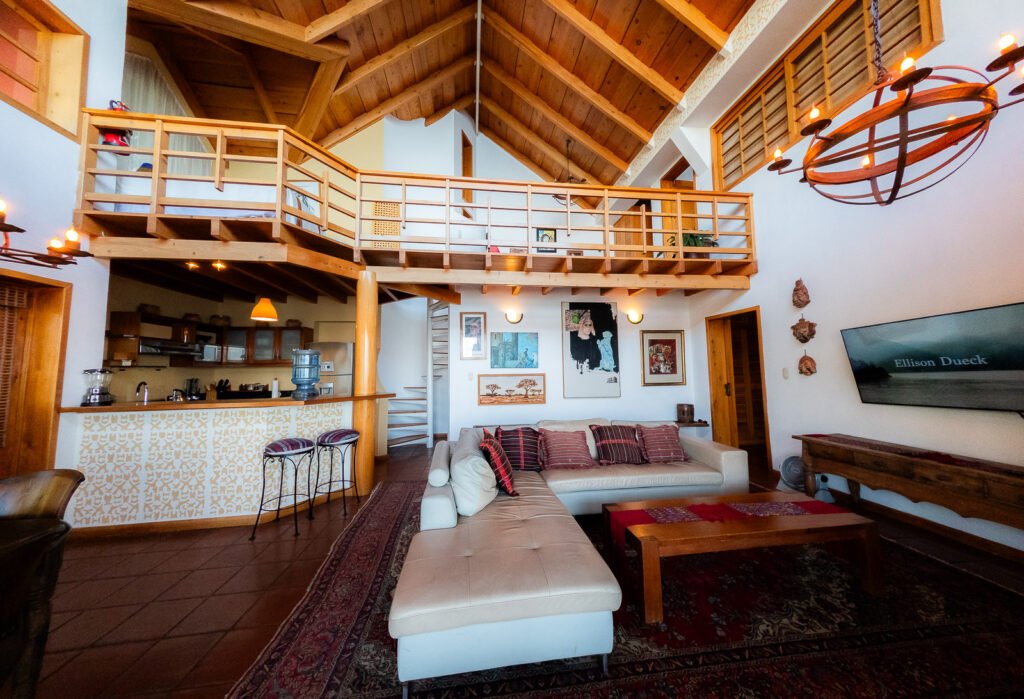 Villa Mirador: Open living space with wood beams, white couch, and balcony overlooking kitchen.