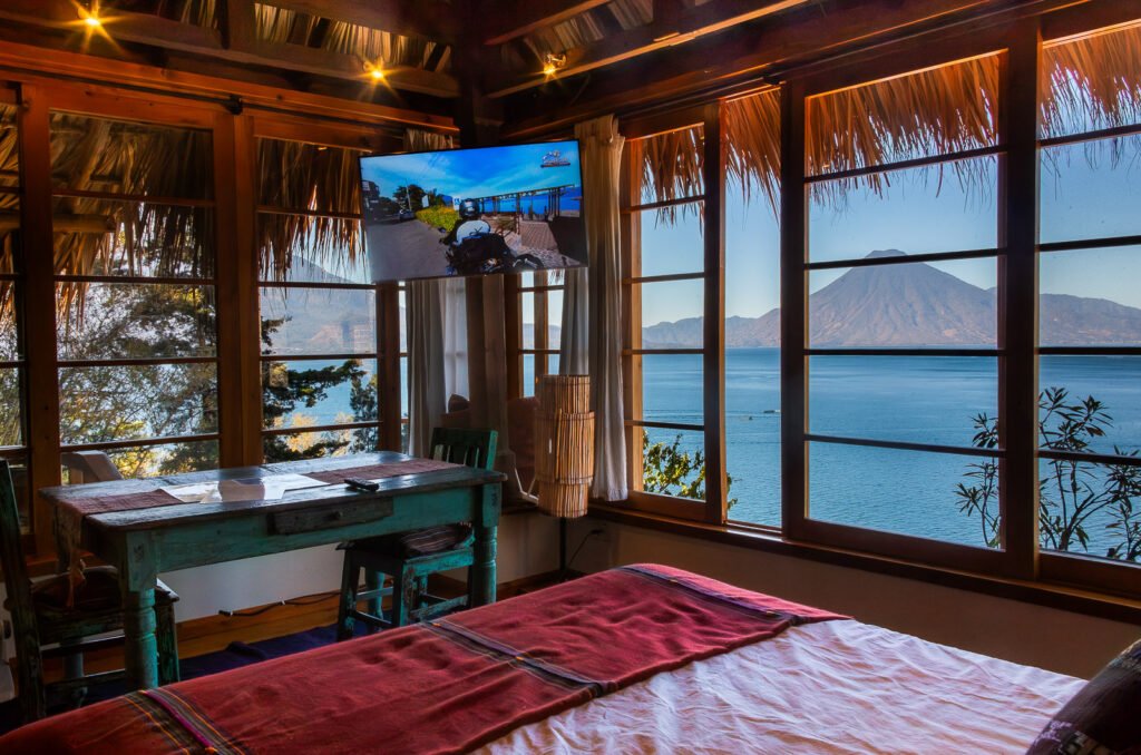 Villa Soluna bedroom with lake view, featuring a rustic table, bed with red blanket, and a large window overlooking the water and mountains.